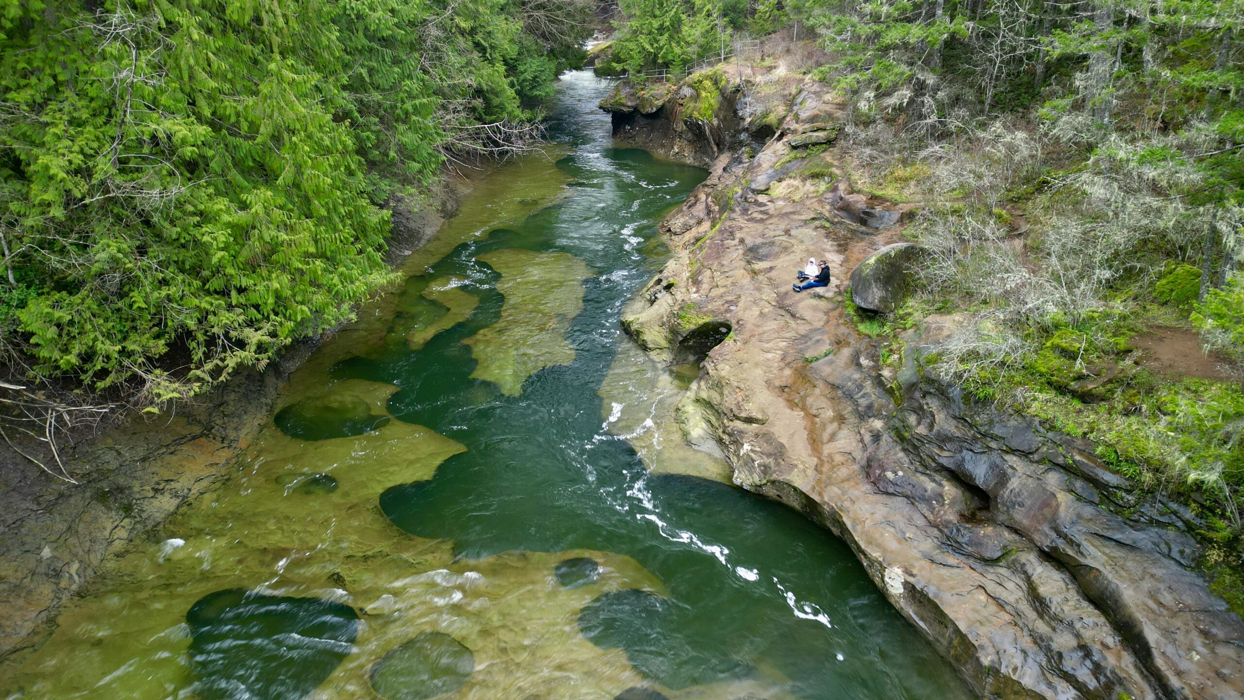 A peaceful aerial view of a river flowing through a lush forest with a person relaxing on rocks.
